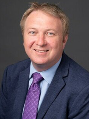 Richard Bates, man with light skin, blonde hair, wearing a dark suit and purple tie